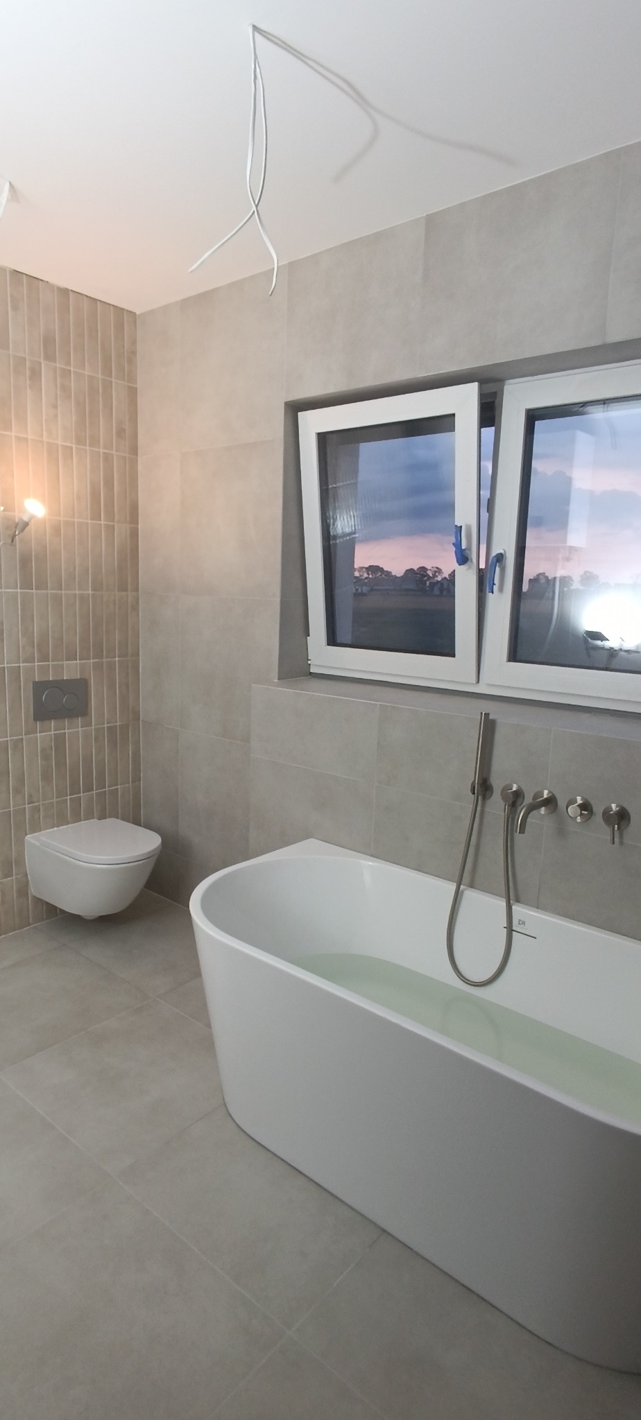 Nowoczesna łazienka z wanną wypełnioną wodą, wiszącą toaletą i oknem z widokiem na zachód słońca. Beżowe płytki na ścianach i podłodze. Widoczne przewody oświetleniowe.