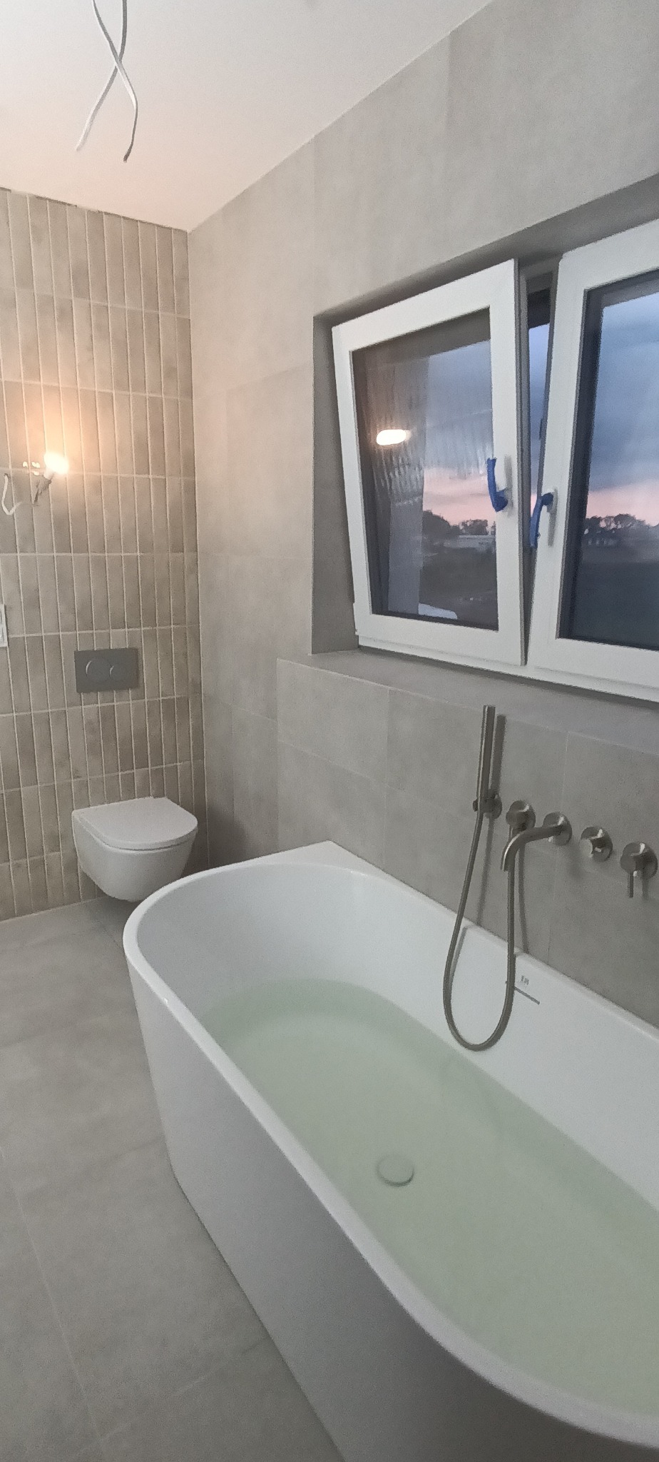 Nowoczesna łazienka w szarościach: wolnostojąca wanna wypełniona wodą, podwieszana toaleta i okno z widokiem na zewnątrz. Minimalistyczny design, płytki imitujące kamień.