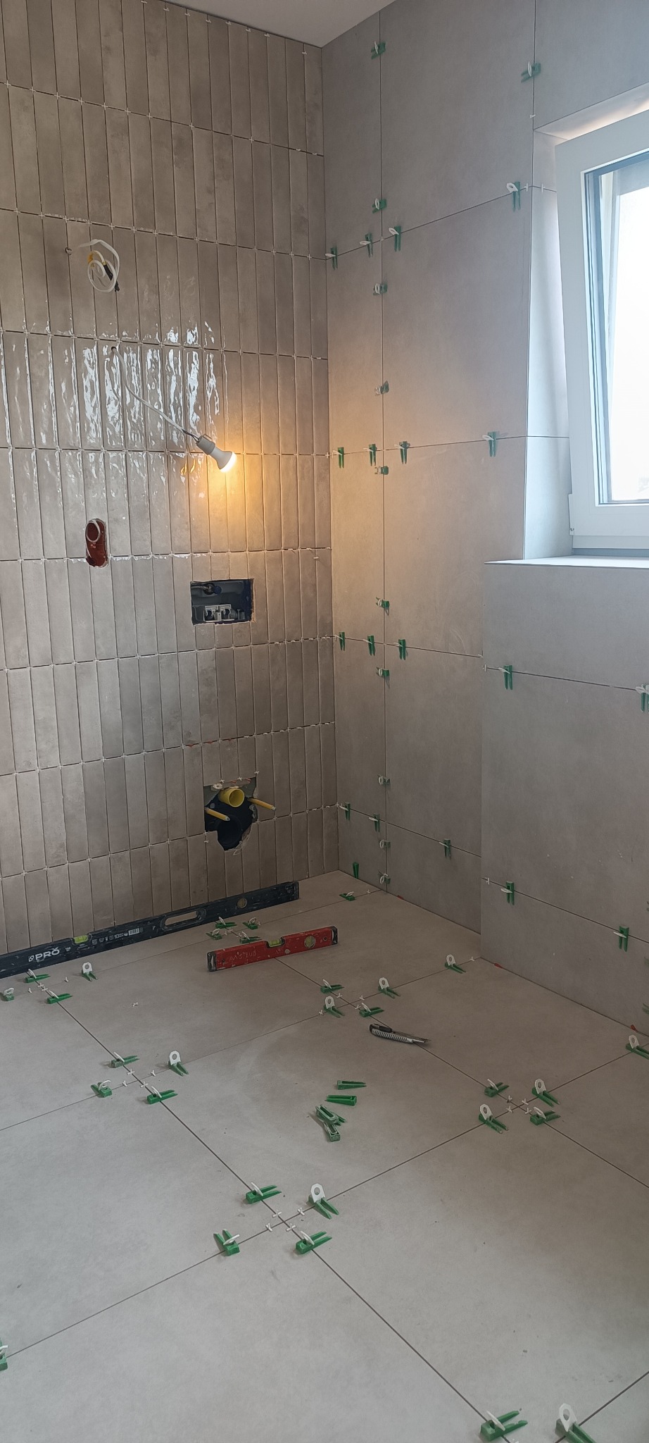 Układanie beżowych płytek w łazience z użyciem poziomicy i klipsów. Widoczne instalacje i okno. Praca w toku, oświetlenie halogenowe.