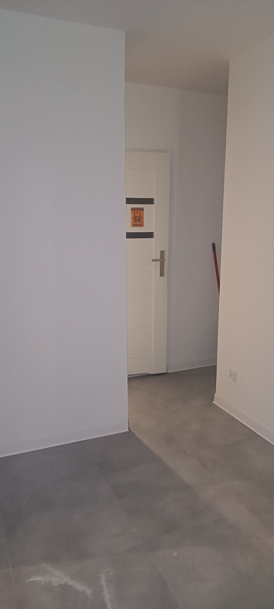 Wnętrze po remoncie: białe ściany, szara podłoga z płytek, białe drzwi z naklejką ostrzegawczą. Widok z perspektywy podłogi, neutralne oświetlenie.