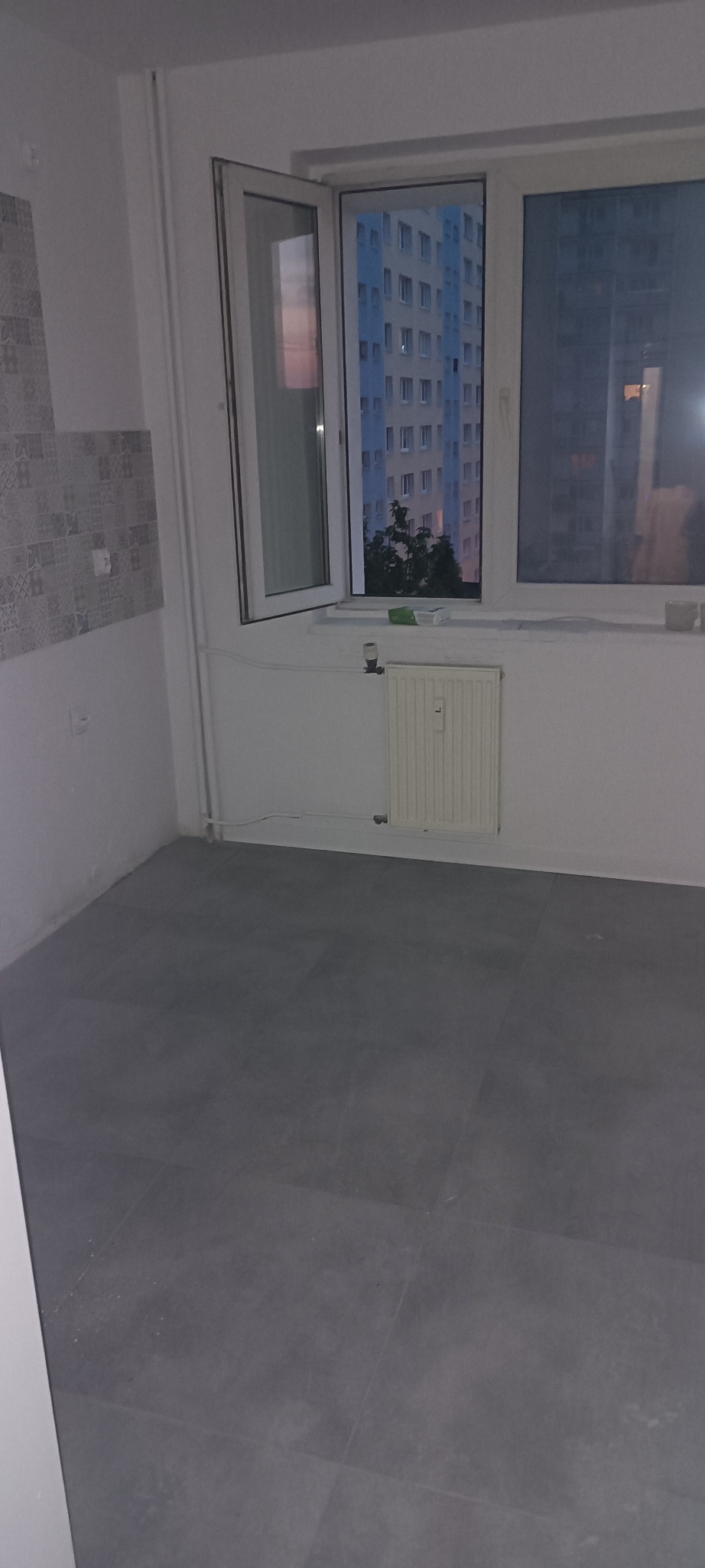 Wykończona szarą płytką podłoga w pustym pokoju z oknem na budynek mieszkalny. Widoczny fragment ściany z dekoracyjną płytką. Grzejnik pod oknem.