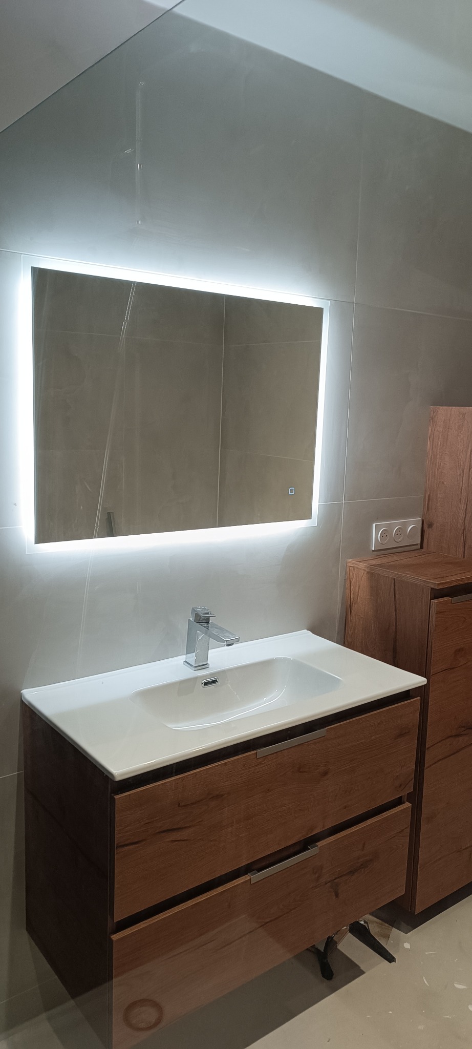 Nowoczesna łazienka: drewniana szafka pod umywalką z białą nablatową umywalką, chromowana bateria i podświetlane lustro na szarej ścianie z płytek.