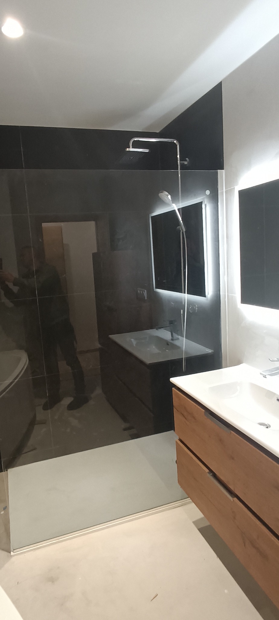 Nowoczesna łazienka z czarnymi płytkami, szklaną kabiną prysznicową i drewnianą szafką pod umywalką. Minimalistyczny design z oświetleniem LED.