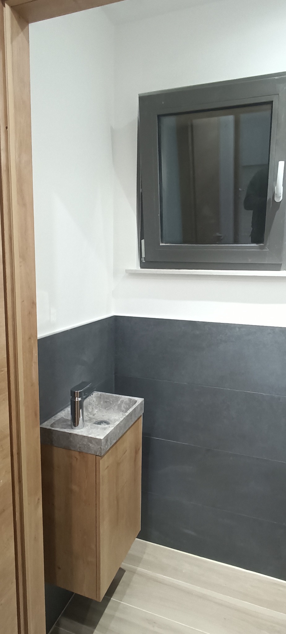 Nowoczesna łazienka z szarą armaturą i drewnianą szafką pod umywalką. Minimalistyczny design, szare płytki na ścianie, okno z ciemną ramą.