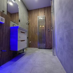 Smart Remonty Wykończenia pod klucz - Nowoczesna łazienka z drewnianą ścianą, szarą płytką i podświetlaną szafką umywalkową. Kabina prysznicowa ze szklanymi drzwiami. Widoczne elementy instalacji.