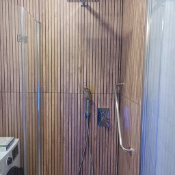 Smart Remonty Wykończenia pod klucz - Nowoczesna kabina prysznicowa z deszczownicą i panelami ściennymi imitującymi drewno. Widoczny uchwyt i bateria. Częściowo widoczna pralka.