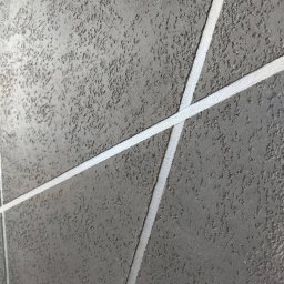 Smart Remonty Wykończenia pod klucz - Szara ściana z dekoracyjną strukturą i białymi, ukośnymi pasami tworzącymi geometryczny wzór. Efektowna faktura i nowoczesny design.