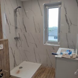 Smart Remonty Wykończenia pod klucz - Wykończona łazienka z kabiną prysznicową, umywalką z szafką i oknem. Płytki imitujące marmur na ścianach i drewnopodobne na podłodze. Widoczny wałek malarski.