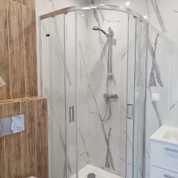 Smart Remonty Wykończenia pod klucz - Nowoczesna kabina prysznicowa z marmurowymi płytkami i drewnopodobnymi akcentami. Widoczna armatura i biała umywalka z szafką. Minimalistyczny design.