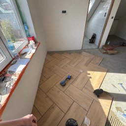 Smart Remonty Wykończenia pod klucz - Układanie płytek podłogowych w jodełkę w łazience z widocznym wejściem do kabiny prysznicowej. Na podłodze narzędzia glazurnicze i resztki kleju.