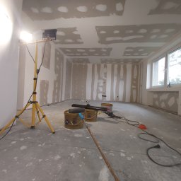 Smart Remonty Wykończenia pod klucz - Pokój w trakcie szpachlowania: ściany z płyt gipsowych, widoczne łączenia, lampa budowlana, wiadra z masą szpachlową i narzędzia malarskie na podłodze.