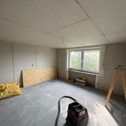 Smart Remonty Wykończenia pod klucz - Pokój w trakcie montażu płyt kartonowo-gipsowych na ścianach i suficie. Widoczny odkurzacz przemysłowy, drabina i materiały izolacyjne. Okno z widokiem na zieleń.