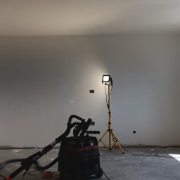 Smart Remonty Wykończenia pod klucz - Wnętrze pokoju po szpachlowaniu ścian, przygotowane do malowania. Widoczny sprzęt budowlany i lampa oświetlająca. Okno z widokiem na zieleń.