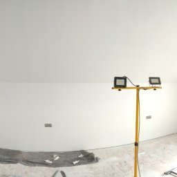 Smart Remonty Wykończenia pod klucz - Poddasze po malowaniu na biało, widoczne okno, lampa budowlana i przygotowane podłoże. Świeżo pomalowane ściany i sufit, gładkie powierzchnie.