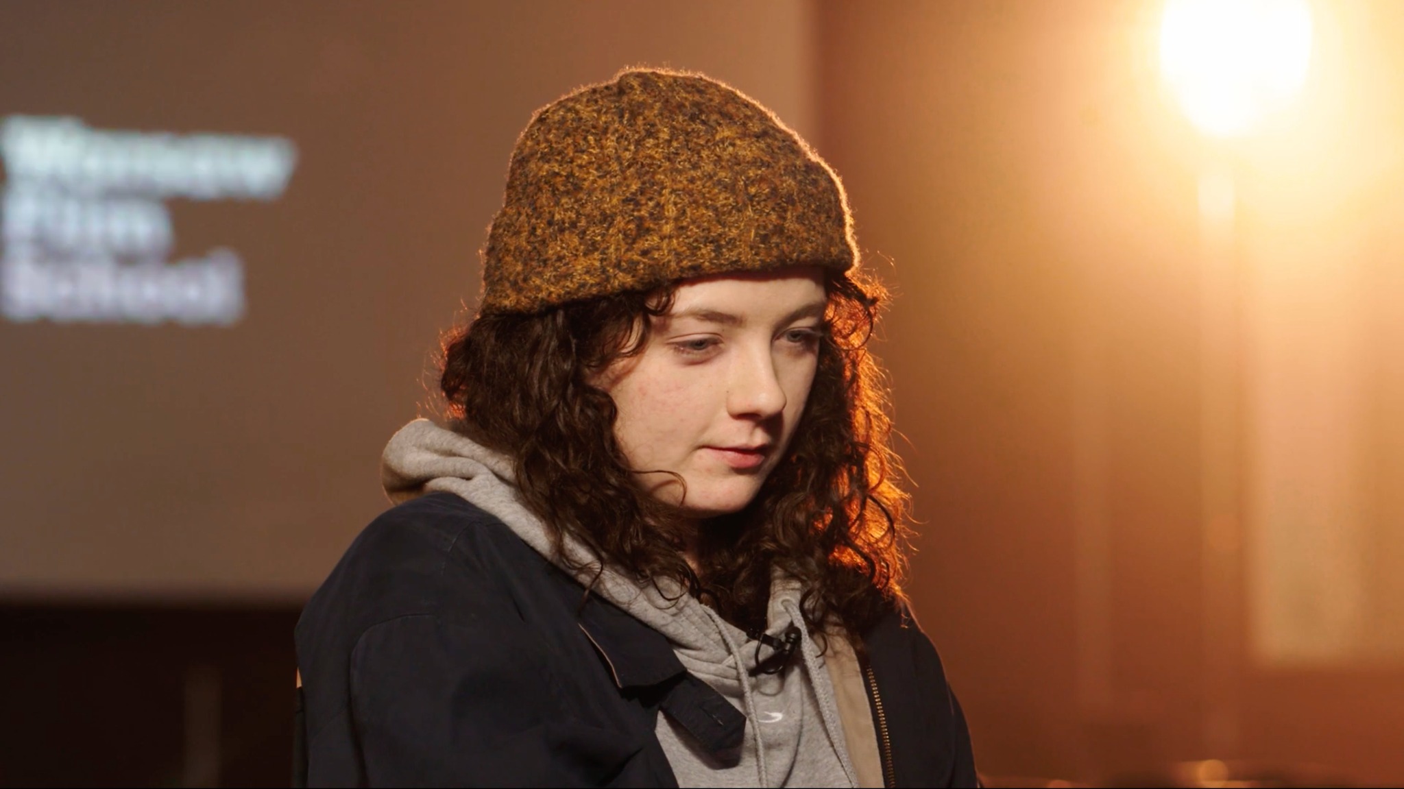 Młoda kobieta w wełnianej czapce i bluzie z kapturem, oświetlona ciepłym światłem, na tle wyświetlonego napisu 'Warszawska Szkoła Filmowa'.