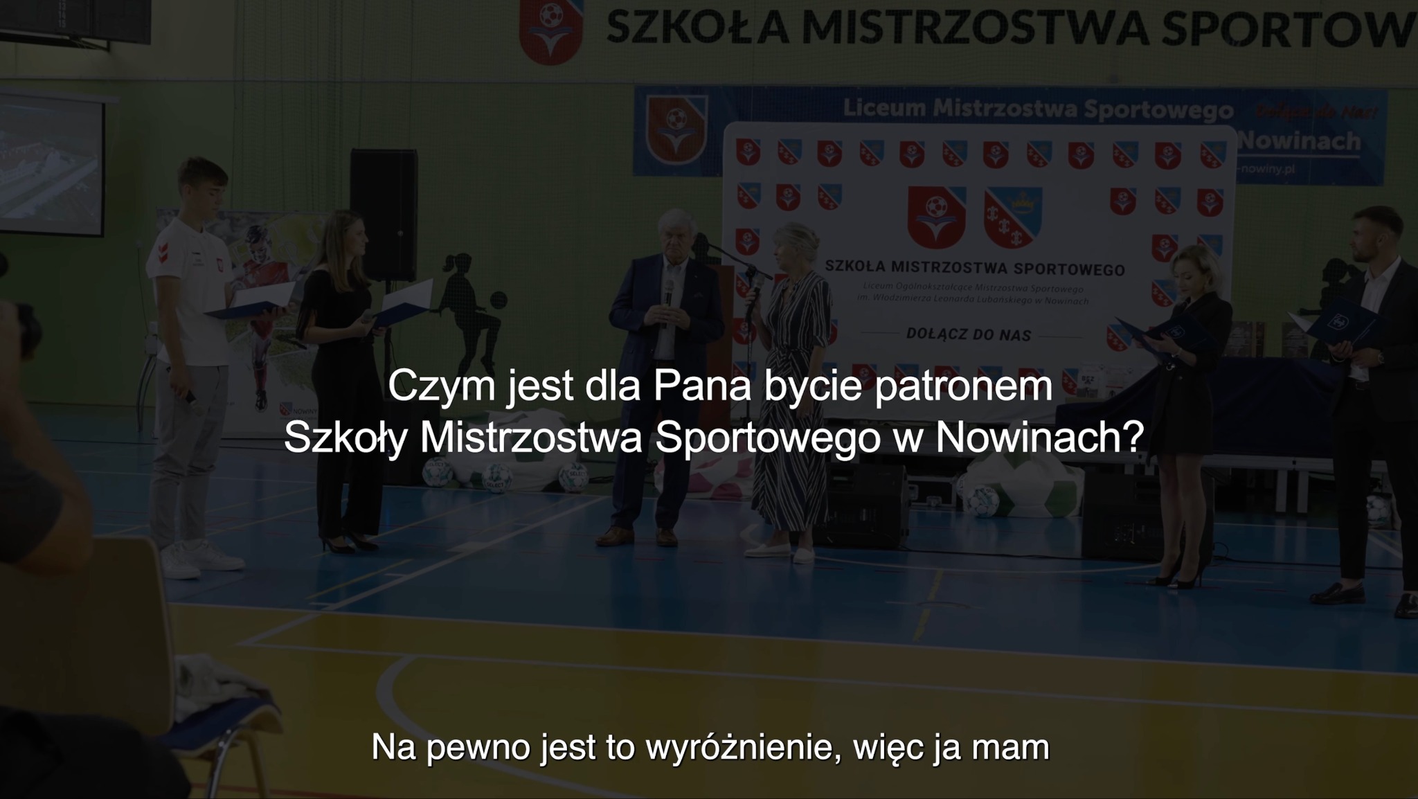 Klatka z reportażu filmowego: Uczniowie i zaproszeni goście podczas uroczystości w Szkole Mistrzostwa Sportowego w Nowinach, na tle baneru z logo szkoły.