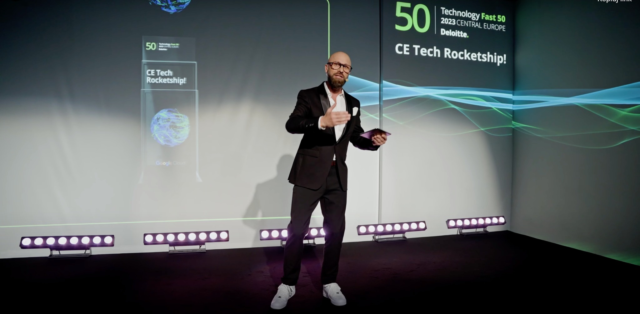 Prezenter w smokingu gestykuluje na scenie podczas gali rozdania nagród Technology Fast 50, na ekranie za nim logo konkursu i grafika z falującymi liniami.