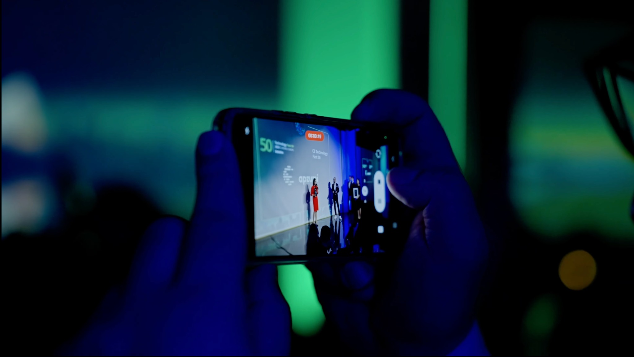 Osoba filmuje smartfonem galę rozdania nagród Fast 50, na ekranie telefonu widoczna scena z prezenterką i laureatami na tle logotypów firm.
