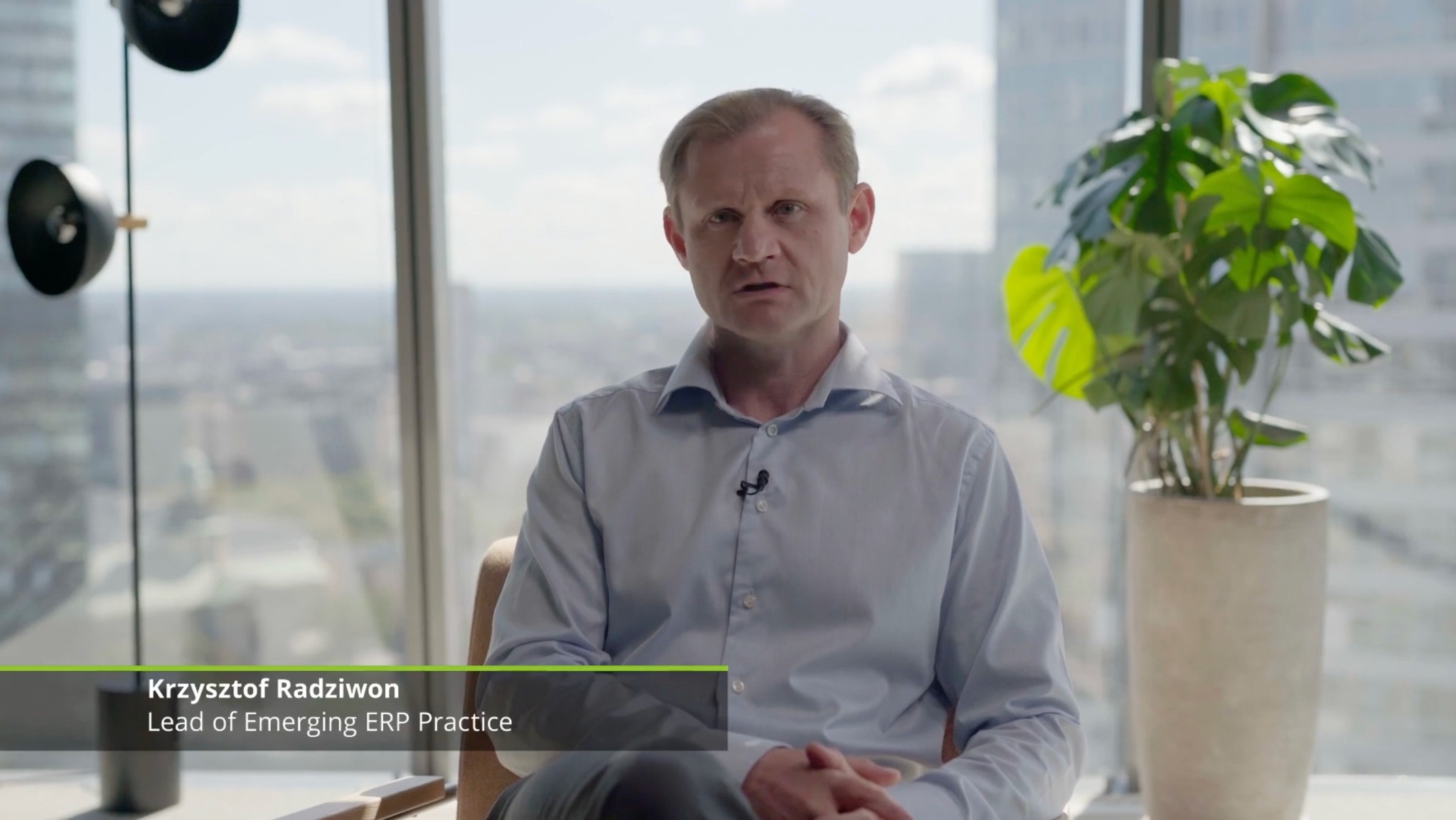 Kadr z filmu promocyjnego: Mężczyzna w błękitnej koszuli siedzi na krześle z widokiem na miasto i roślinę w doniczce, prezentuje się przed kamerą.