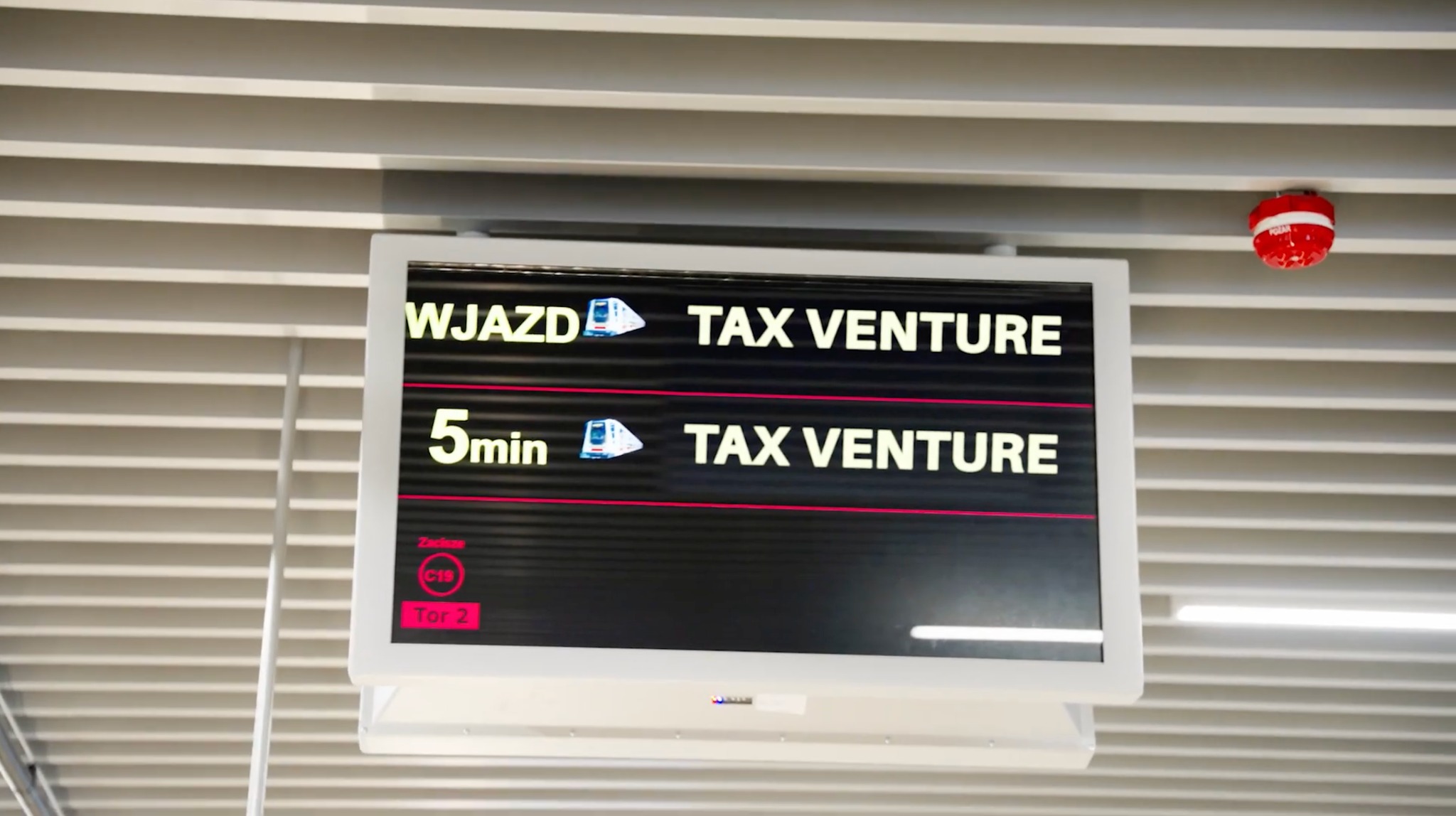 Ekran informacyjny z napisem 'Wjazd' oraz '5min' i logotypem 'TAX Venture' na tle sufitu z żaluzjami i czerwonym czujnikiem pożarowym.