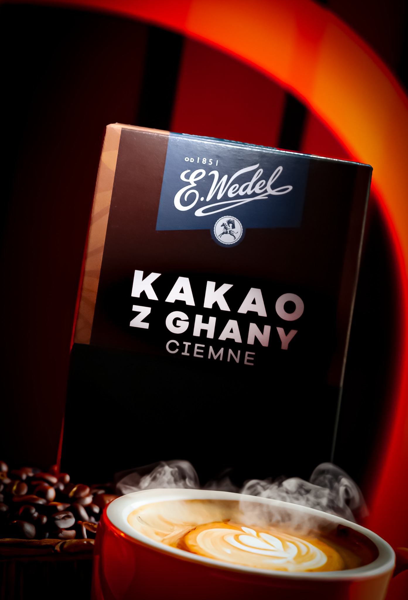 Pudełko kakao E.Wedel z Ghany Ciemne, otoczone ziarnami kawy i parującą filiżanką latte art na czerwonym tle.