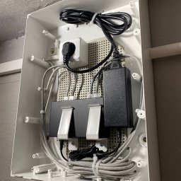 FH-ELEKTRO - Wnętrze białej skrzynki z zamontowanym rozdzielaczem sieciowym, zasilaczem i okablowaniem. Widoczne gniazdo z wtyczką i przewody w szarych osłonach.