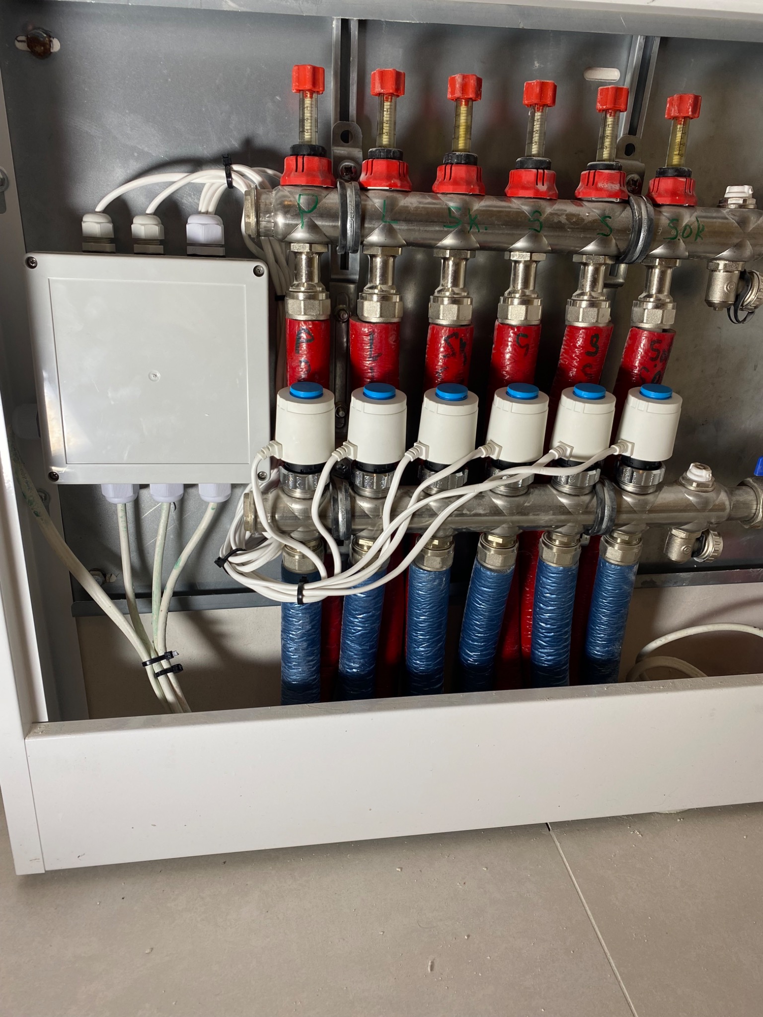 Rozdzielacz ogrzewania podłogowego w szafce, z widocznymi zaworami, rurami w izolacji i przewodami elektrycznymi. Elementy instalacji w kolorach czerwonym, niebieskim i białym.