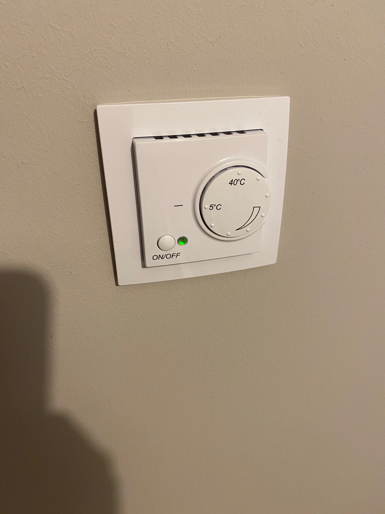 Biały termostat pokojowy z zieloną diodą ON/OFF, zamontowany na beżowej ścianie. Widoczna skala temperatury od 5 do 40 stopni Celsjusza.