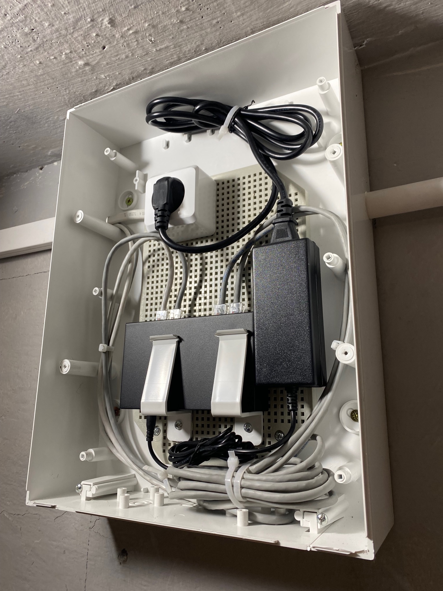 Wnętrze białej skrzynki z zamontowanym rozdzielaczem sieciowym, zasilaczem i okablowaniem. Widoczne gniazdo z wtyczką i przewody w szarych osłonach.
