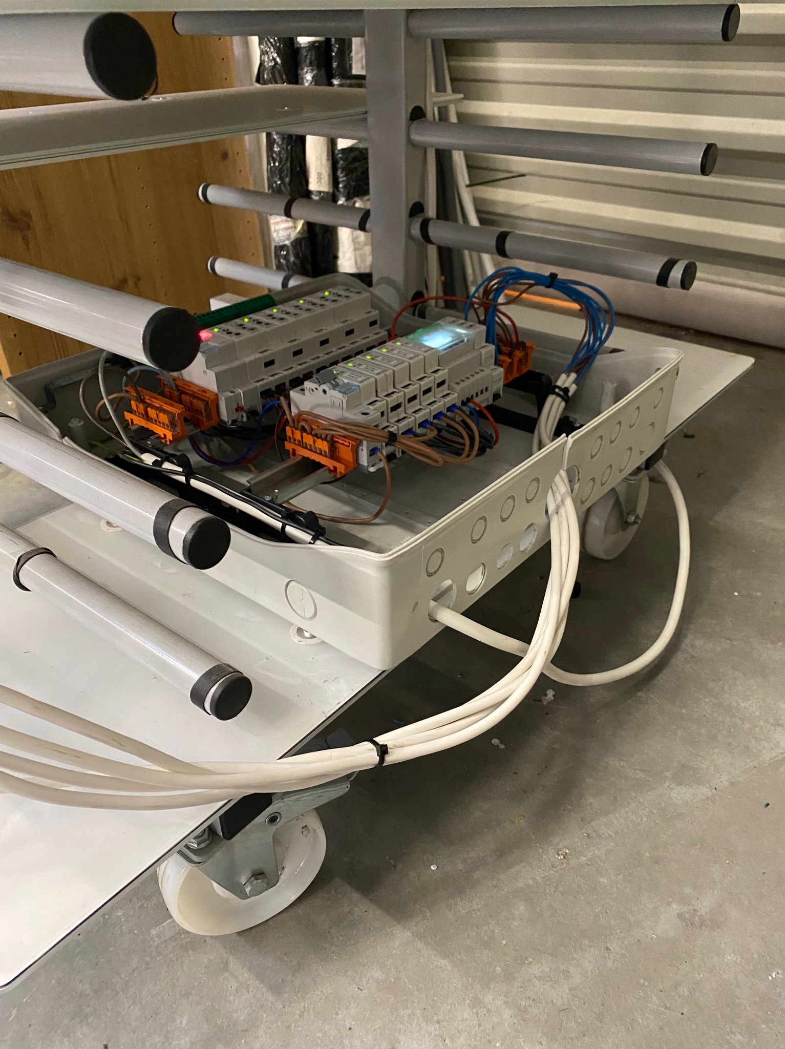 Mobilna skrzynka elektryczna z przewodami i sterownikami, na kółkach, w warsztacie. Widoczne elementy instalacji i okablowanie.