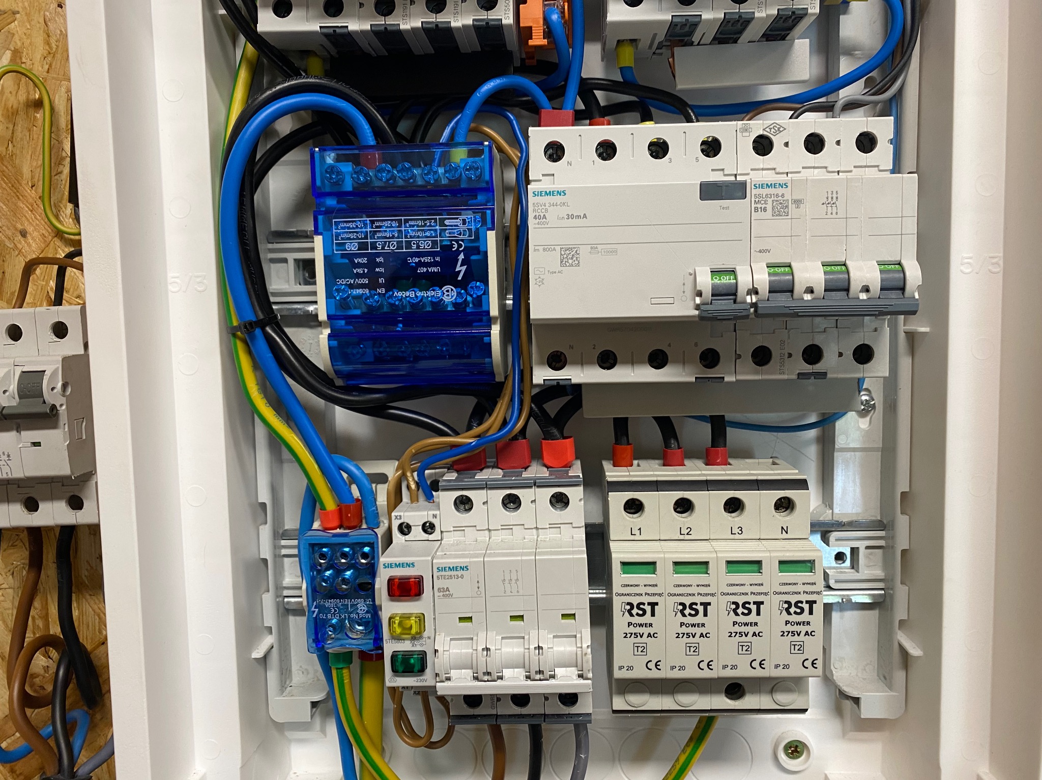 Rozdzielnica elektryczna z zabezpieczeniami przepięciowymi i wyłącznikami różnicowoprądowymi marki Siemens, z widocznymi przewodami w kolorach niebieskim, żółto-zielonym i czarnym.