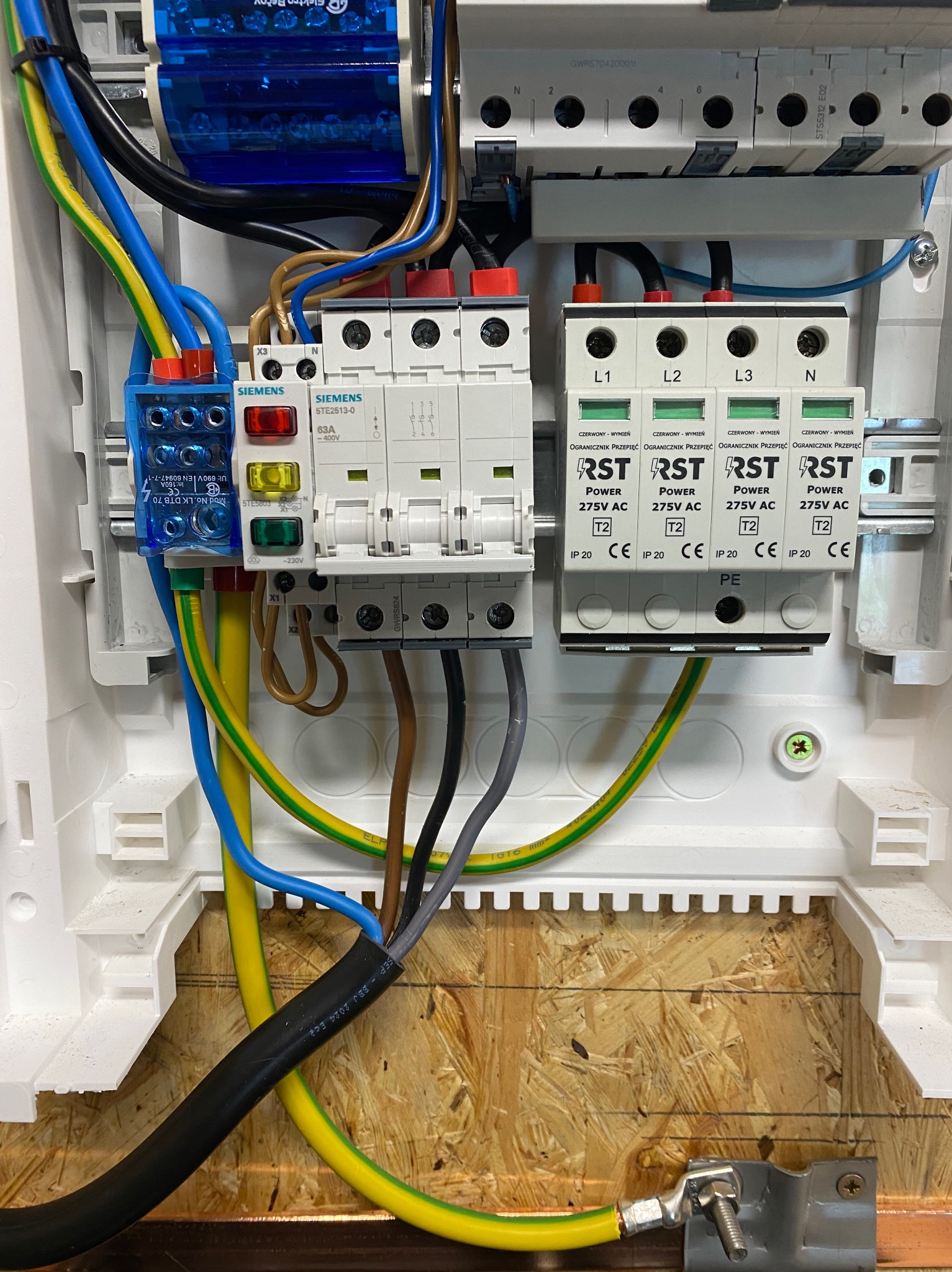 Rozdzielnica elektryczna z widocznymi bezpiecznikami, przewodami w kolorach: żółto-zielonym, niebieskim i brązowym. Elementy marki Siemens i RST.