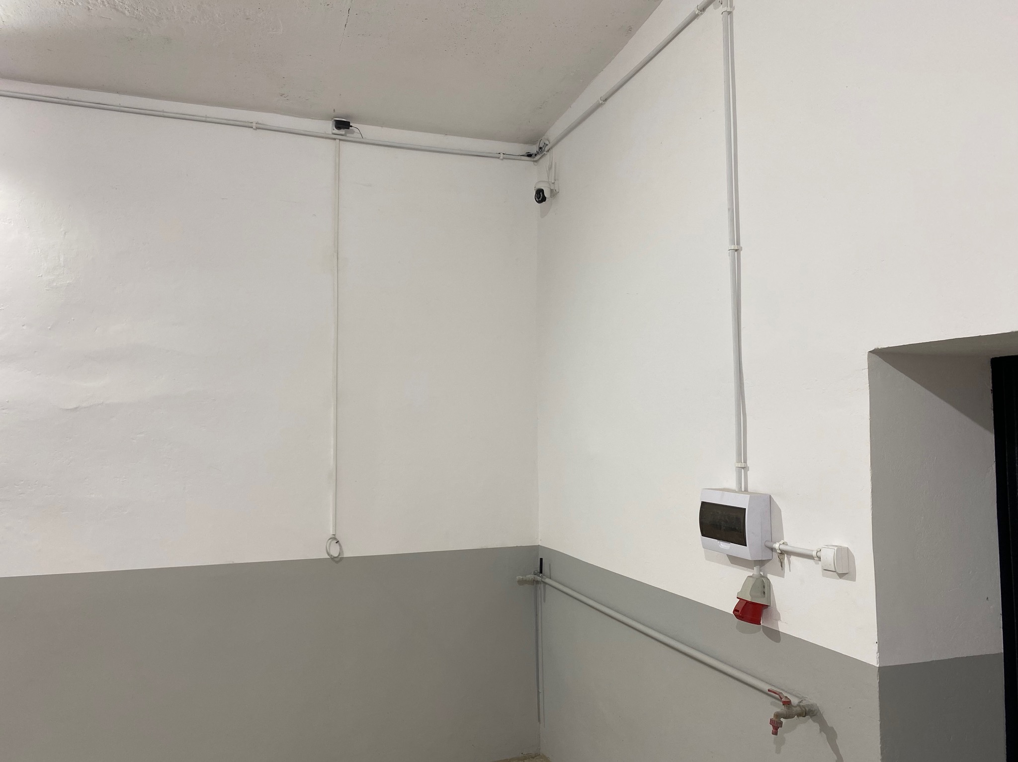 Białe ściany z szarą lamperią, instalacje elektryczne w rurkach, puszka elektryczna z gniazdem i kamera przemysłowa w rogu pomieszczenia. Minimalistyczne wykończenie.