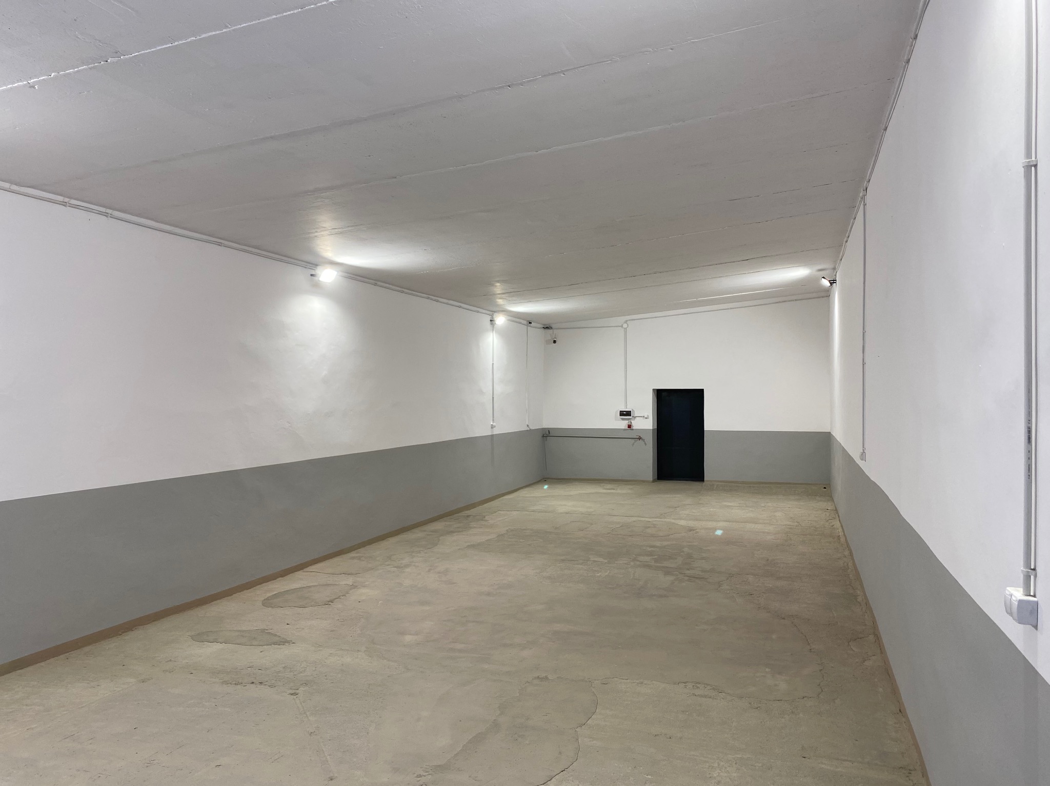 Puste, odnowione pomieszczenie z szarą lamperią i nową instalacją elektryczną. Widoczne kable w białych listwach i równomiernie rozmieszczone lampy sufitowe.