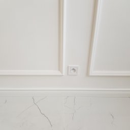 ALL-TRADE Sp. z o.o. - Biała ściana z dekoracyjnymi listwami, gniazdkiem elektrycznym i marmurową podłogą z szarymi żyłami.