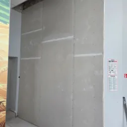 Ściana z płyt kartonowo-gipsowych, widoczne łączenia płyt wypełnione masą szpachlową. Do ściany przymocowane instrukcje bezpieczeństwa.