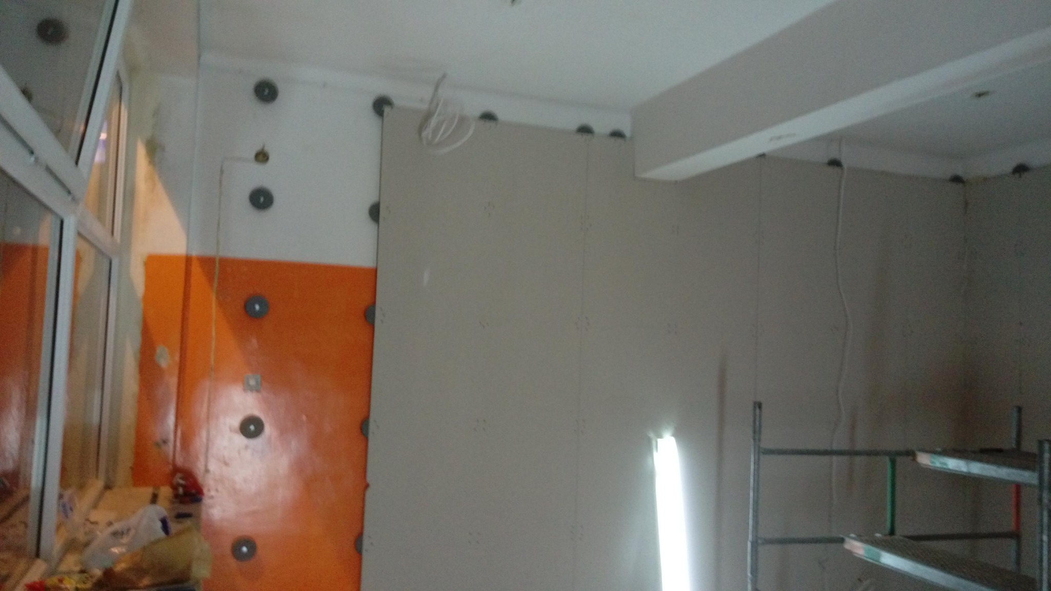 Montaż płyt na ścianie z widocznym profilem i izolacją, fragment rusztowania w rogu pokoju, przewody zwisające z sufitu.