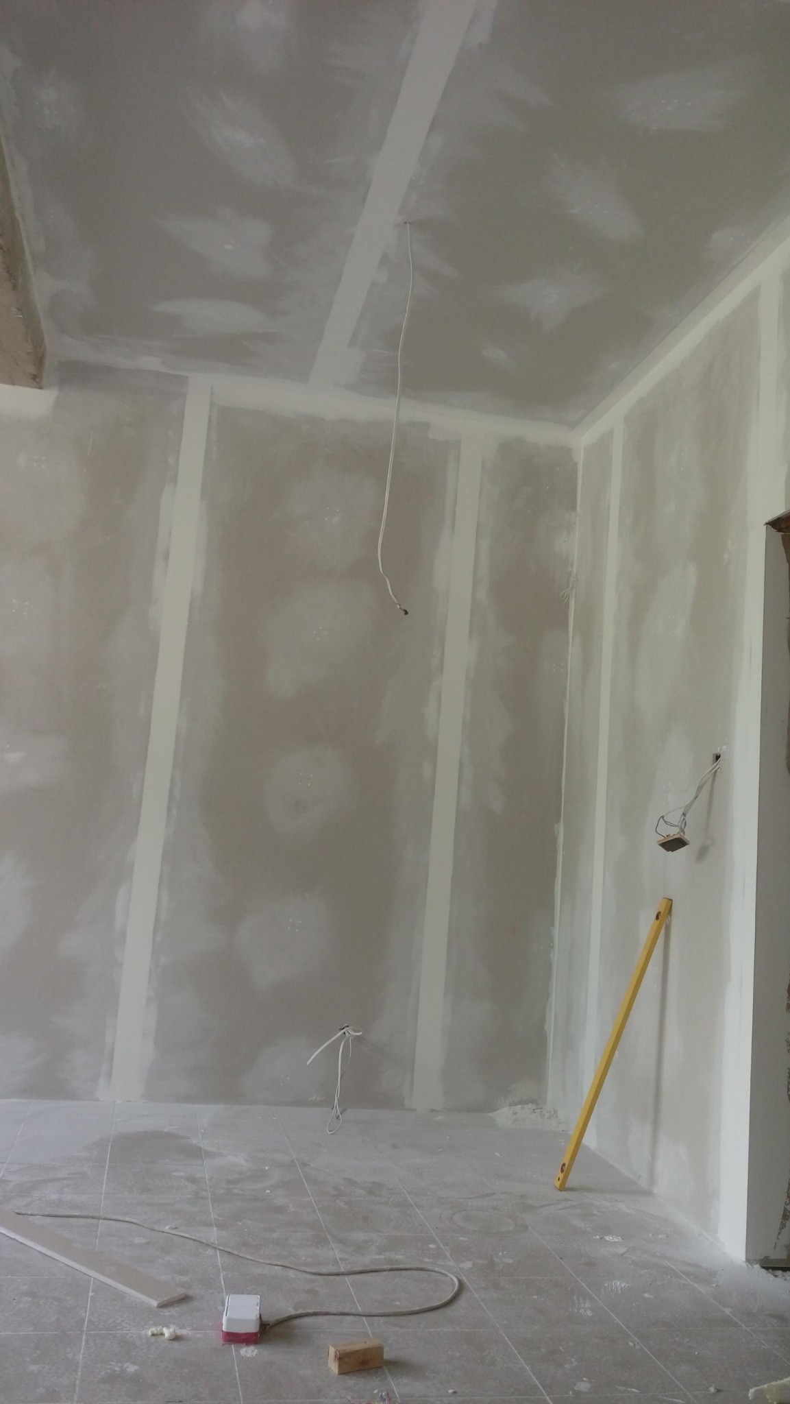 Wnętrze w trakcie prac, ściany i sufit pokryte wstępną warstwą gładzi na płytach gipsowo-kartonowych, widoczne kable elektryczne i poziomica oparta o ścianę.