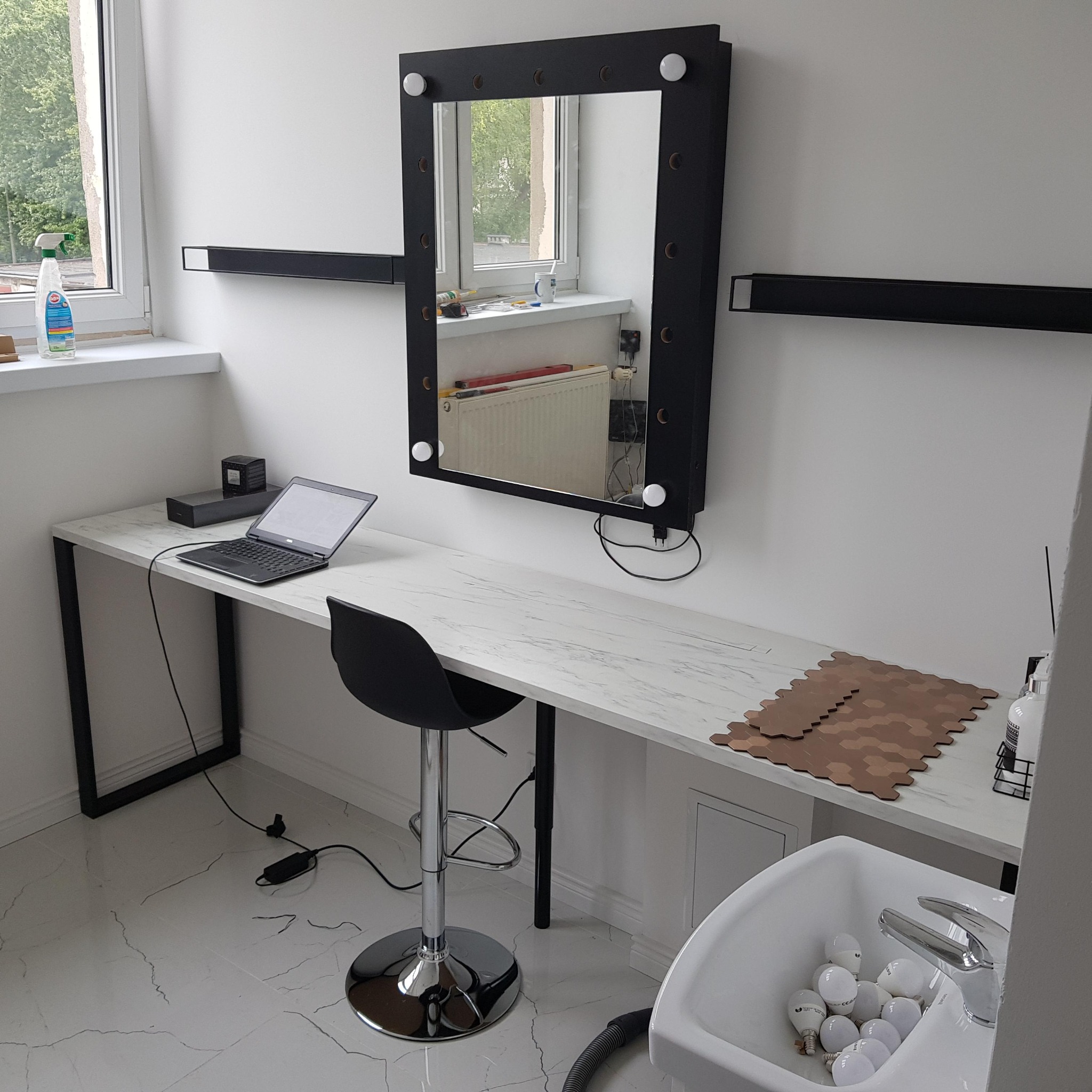 Wnętrze salonu kosmetycznego w trakcie urządzania: toaletka z marmurowym blatem, podświetlane lustro w czarnej ramie, czarne półki, brązowe płytki heksagonalne, umywalka z żarówkami.