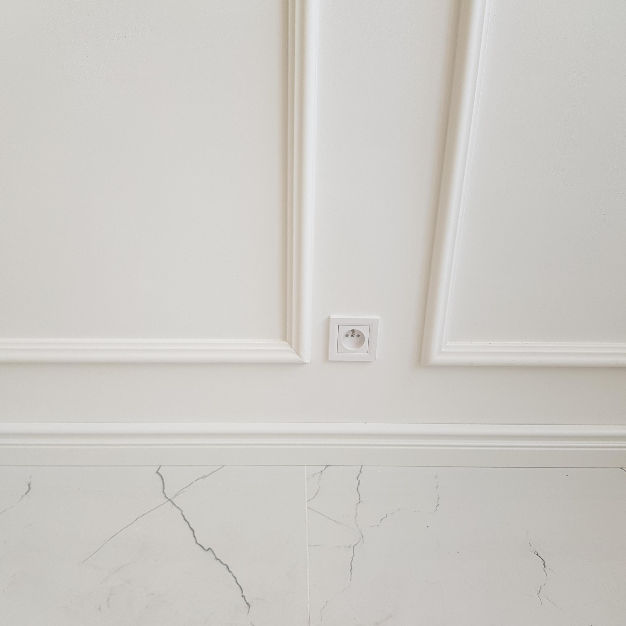 Biała ściana z dekoracyjnymi listwami, gniazdkiem elektrycznym i marmurową podłogą z szarymi żyłami.