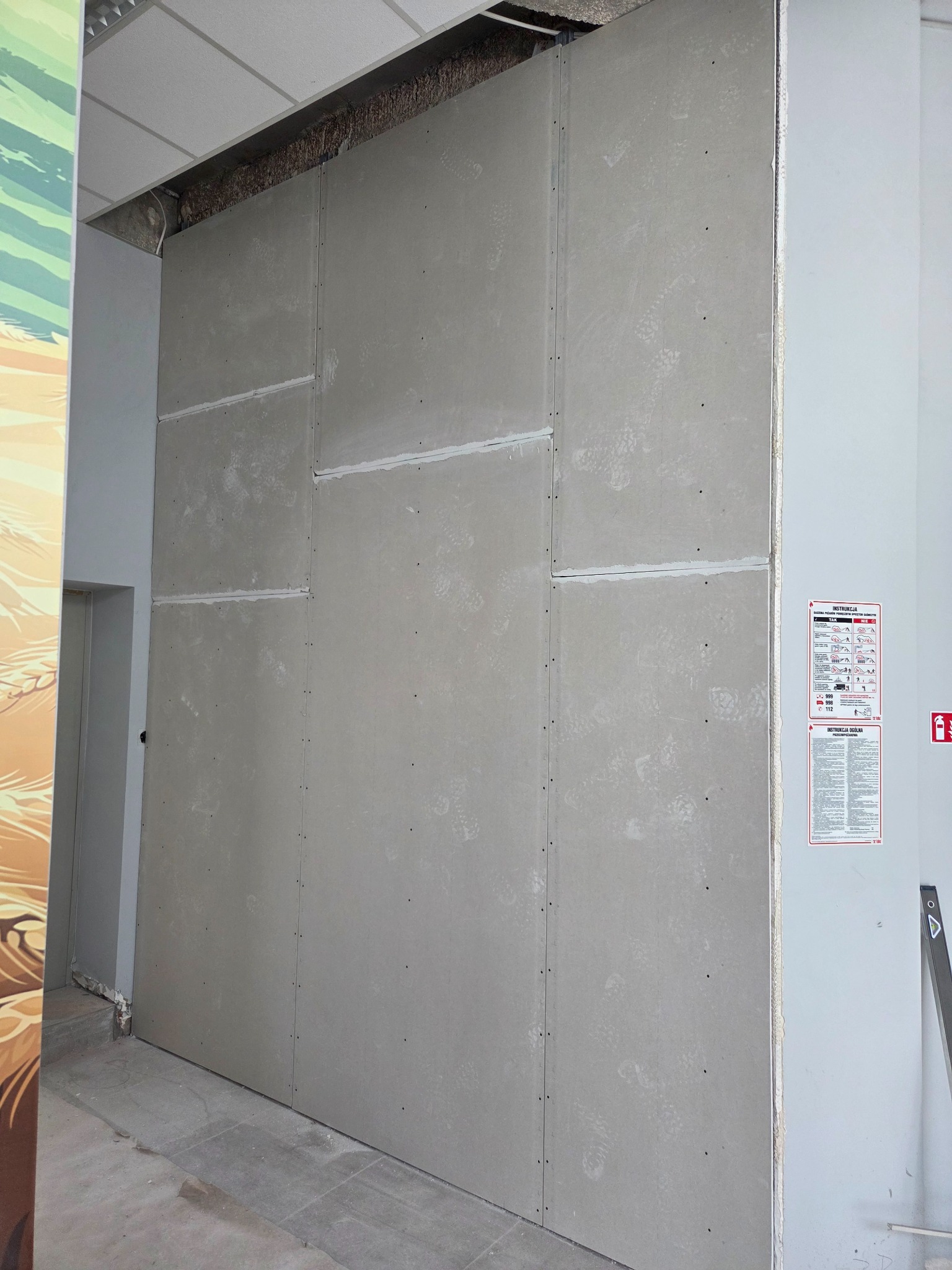 Ściana z płyt kartonowo-gipsowych, widoczne łączenia płyt wypełnione masą szpachlową. Do ściany przymocowane instrukcje bezpieczeństwa.