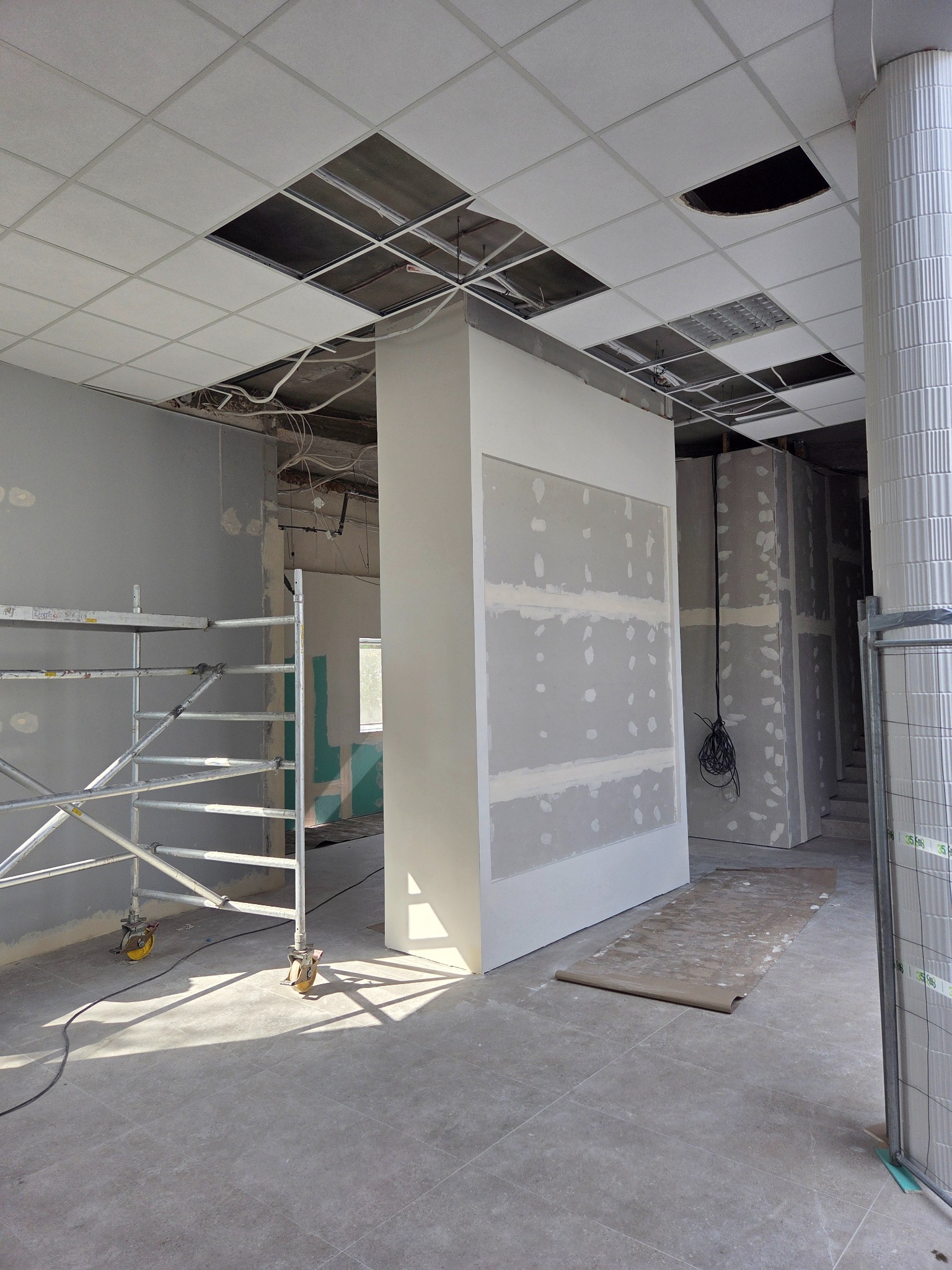 Wnętrze biura w trakcie prac wykończeniowych: ściany z płyt gipsowo-kartonowych, szpachlowane, sufit podwieszany z brakującymi panelami, rusztowanie i zabezpieczenia.