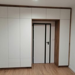 JMM PROJEKT Michał Marciniak - Biała szafa wnękowa z drewnianą ramą okalającą również drzwi wejściowe, tworząc spójny wizualnie układ zabudowy.