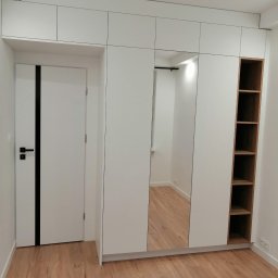 JMM PROJEKT Michał Marciniak - Biała szafa wnękowa z lustrem i otwartymi półkami z jasnego drewna, dopasowana do białych drzwi z czarnym akcentem w nowoczesnym wnętrzu z drewnianą podłogą.
