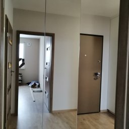 JMM PROJEKT Michał Marciniak - Szafa wnękowa z lustrzanymi drzwiami przesuwnymi w przedpokoju, odbijająca wnętrze mieszkania, widoczna podłoga laminowana i drzwi wejściowe.