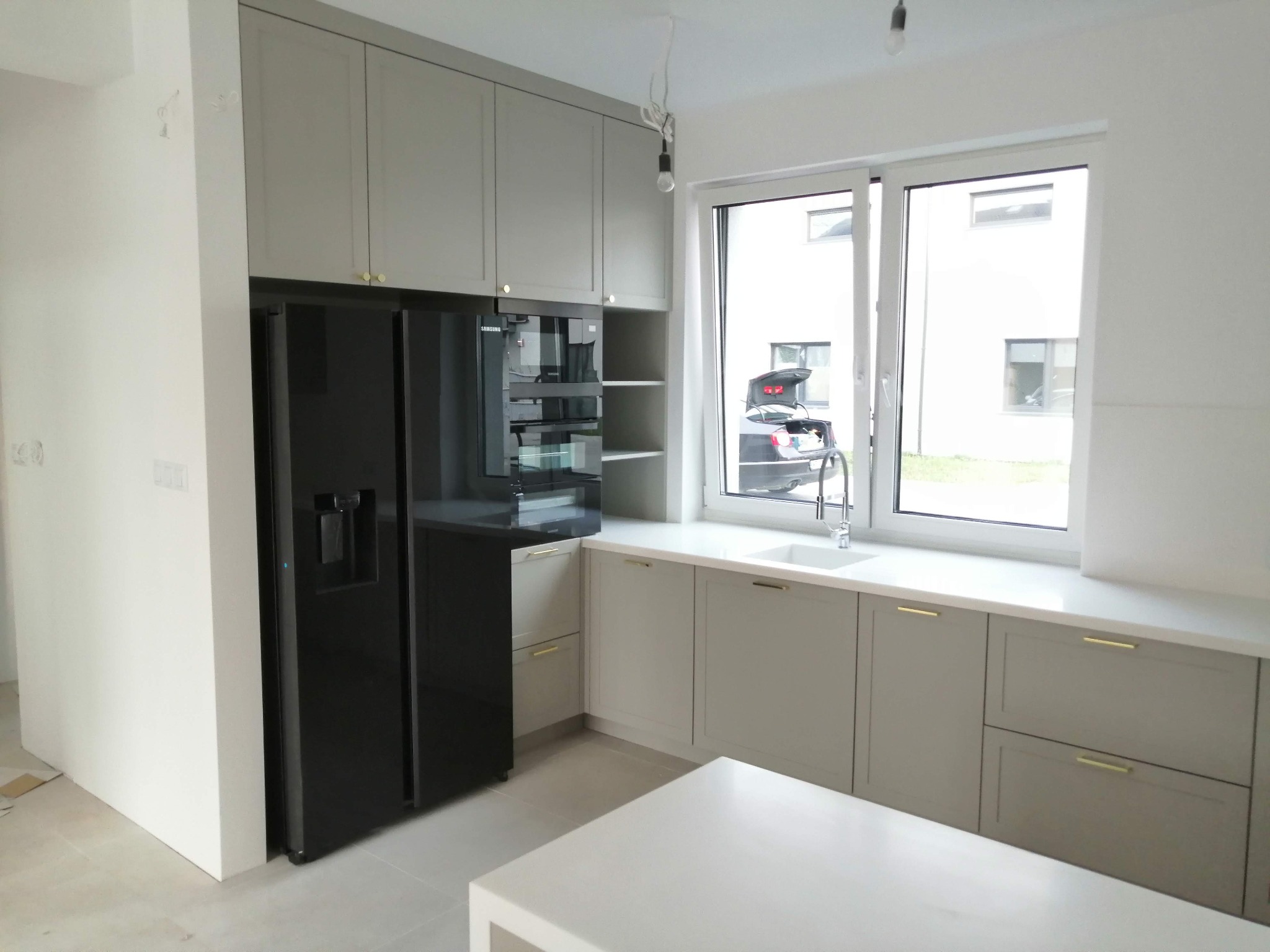 Jasnoszara zabudowa kuchenna z czarną lodówką side-by-side i widokiem na samochód z otwartym bagażnikiem przez okno