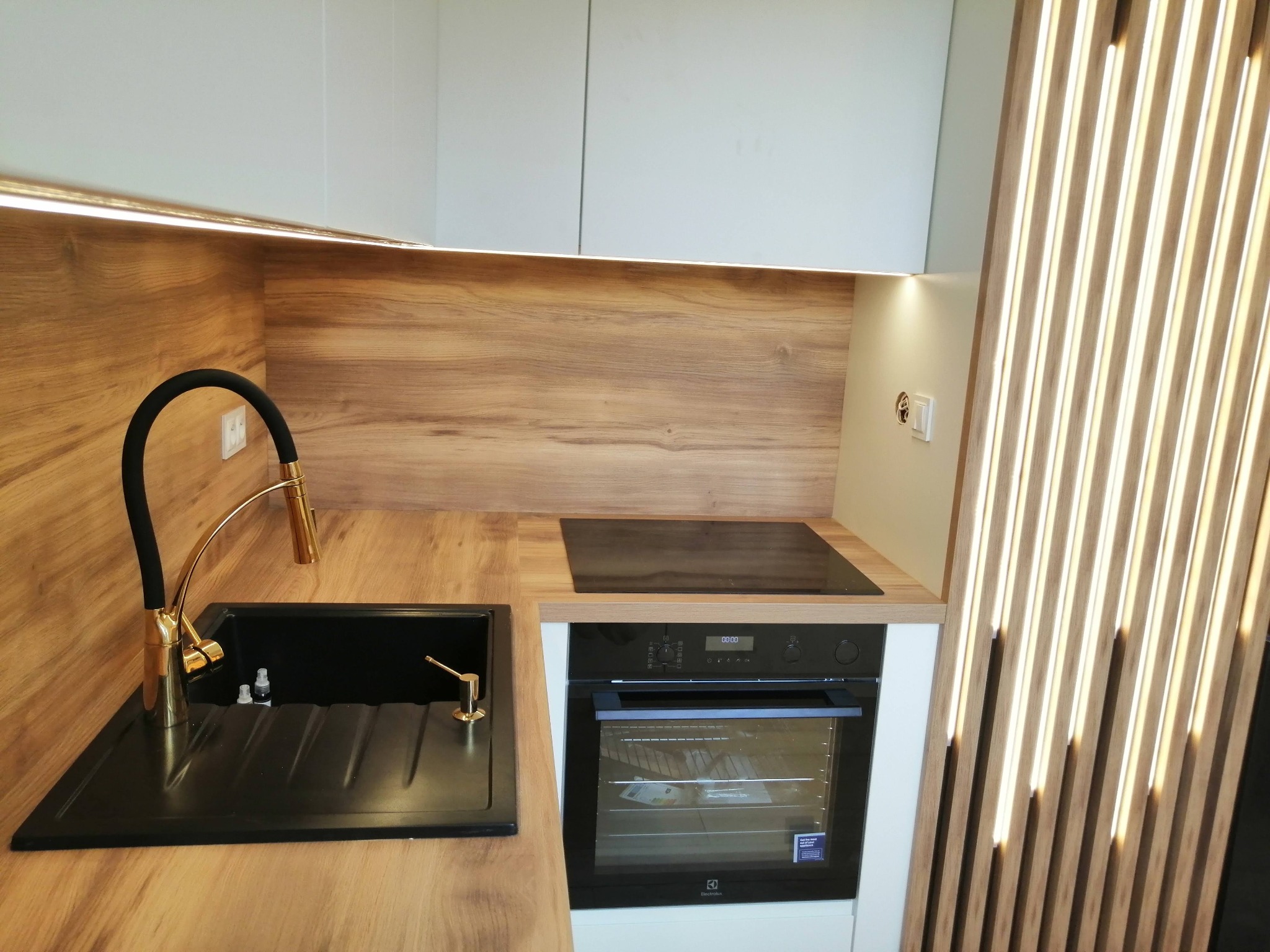 Nowoczesna kuchnia z drewnianymi akcentami: czarny zlew z złotą armaturą, płyta indukcyjna i piekarnik Electrolux w zabudowie, oświetlenie LED podszafkowe i dekoracyjna ścianka z drewnianych listew.