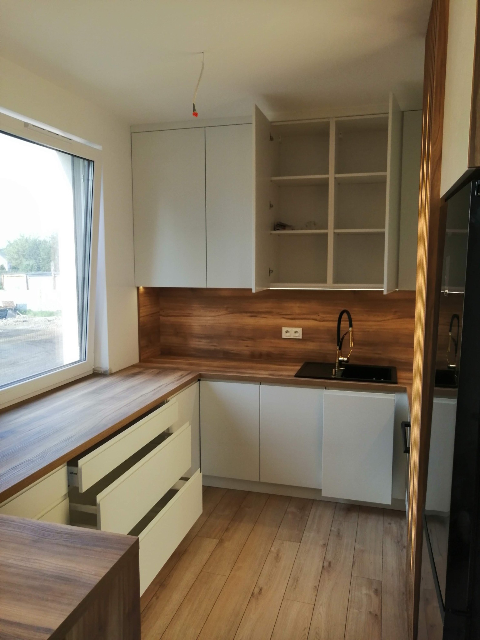 Nowoczesna kuchnia z białymi szafkami, drewnianym blatem i złotą armaturą, z otwartymi szufladami i widocznym okablowaniem na suficie.