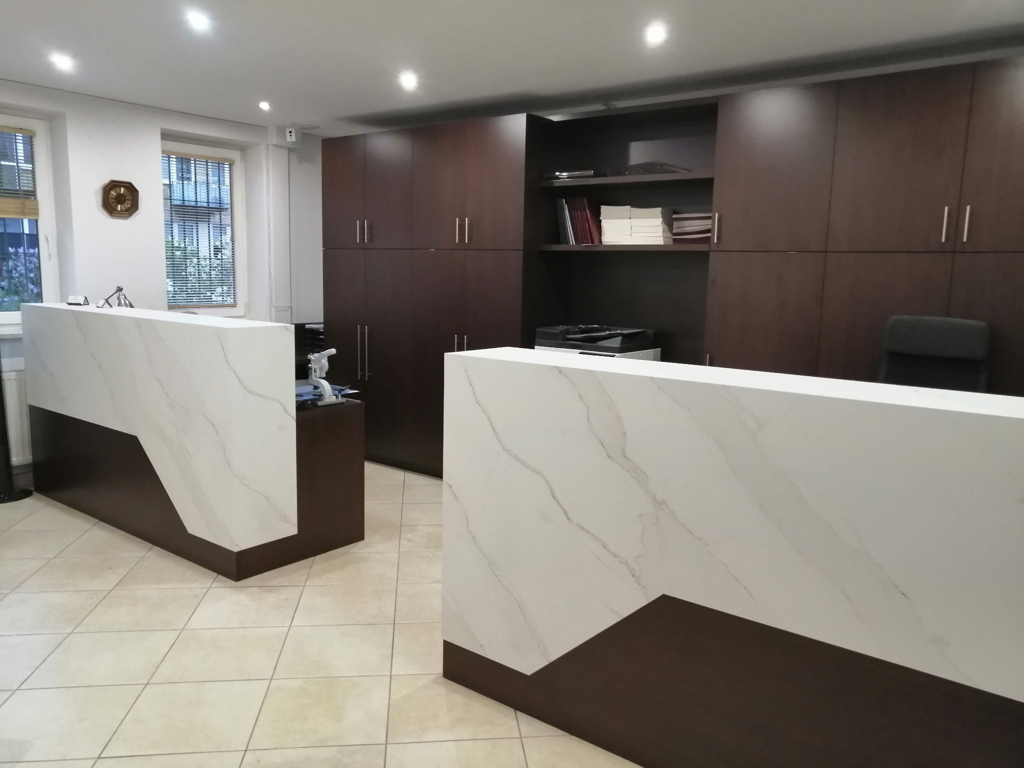 Nowoczesna recepcja z dwoma stanowiskami o marmurowym wykończeniu, ciemnobrązowe szafy w tle, jasna podłoga z płytek.