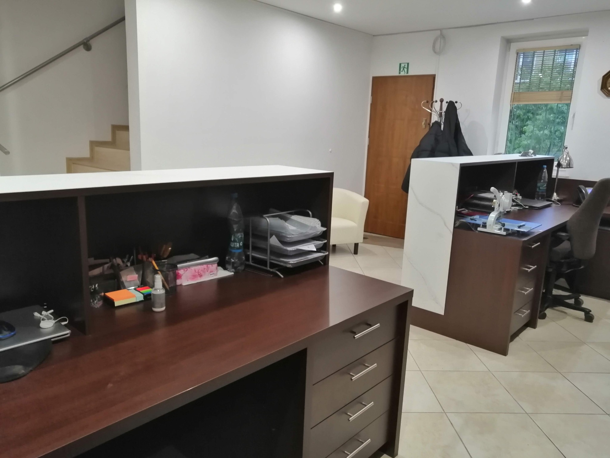 Biuro z dwoma stanowiskami recepcyjnymi z ciemnego drewna i białymi elementami, widoczne schody i krzesło w tle.